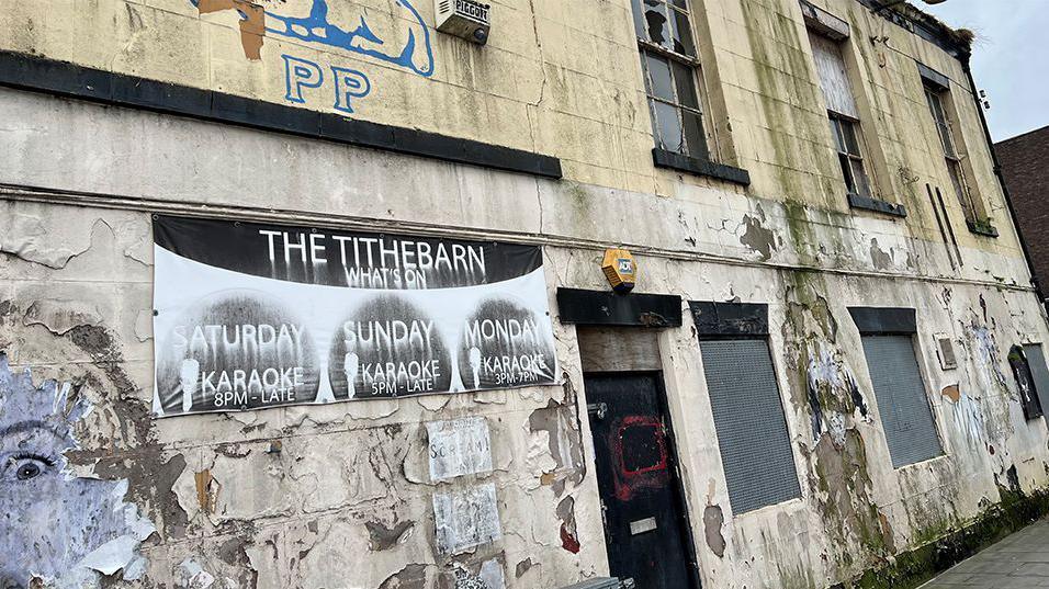 The outside of the Tithebarn pub, a dirty, tired-looking building with peeling white paint, rain damage, boarded up windows and peeling posters