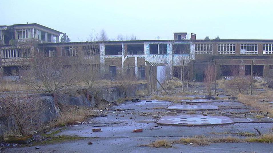 The deserted site of Huncoat power station. It is a long two-storey brick building with a taller section to the left which have been vandalised. To the front are overgrown flat areas with covers on the ground and sprouting weeds