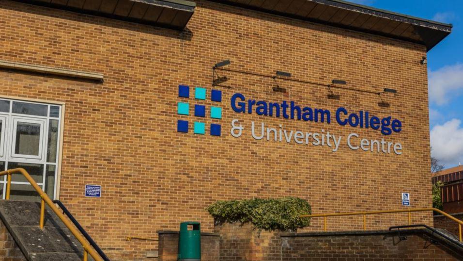 The side of a brick building with large words 'Grantham College & University Centre' written in blue and silver. You can see steps leading up to white doors on the left and a green bin in the middle.