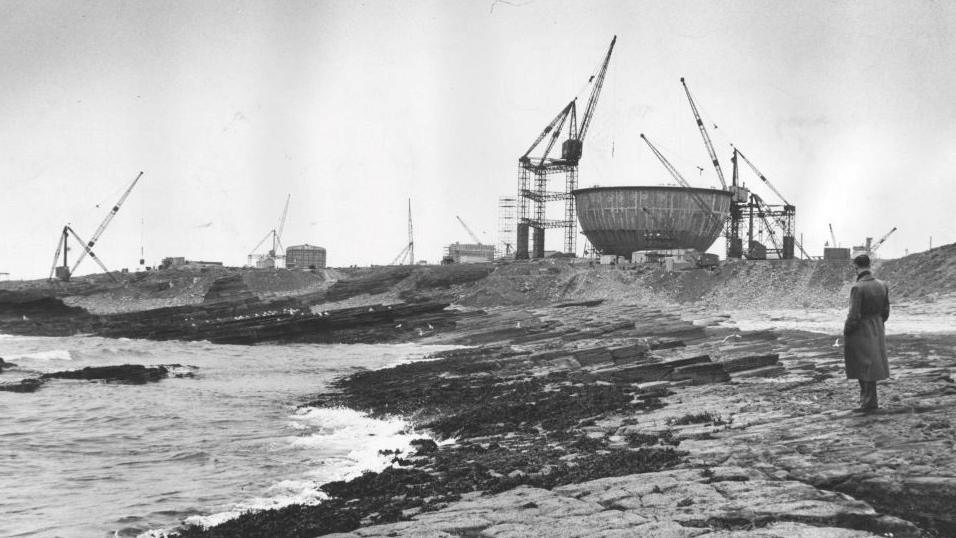 A man stands on a rocky shore and looks towards cranes involved in the construct of Dounreay's facilities, including its famous reactor dome.