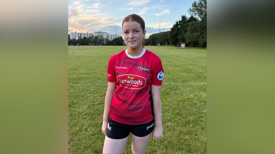 Aoibhinn standing on the pitch, she's wearing a red rugby top that says Armagh on it alongside sponsors. Her red hair is tied up in a ponytail and she is smiling. 