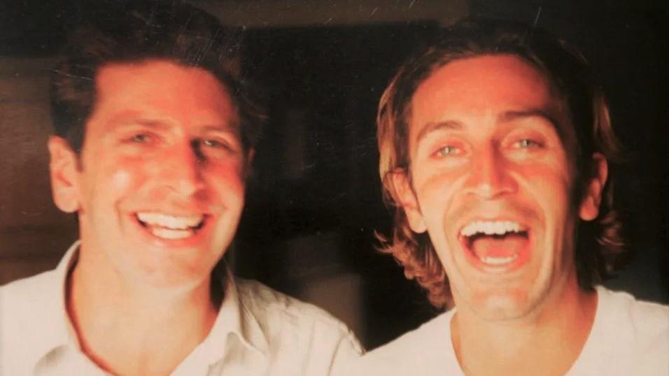 Piers Simon on the left wearing a white collared shirt. He has dark hair, green eyes, and is smiling at the camera. His younger brother Luke is on the right, wearing a white t-shirt and laughing at the camera. He has green eyes, and shoulder-length brown hair with blonde highlights.