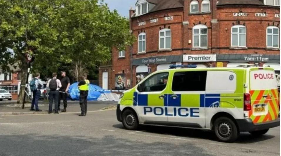 A police van parked in front of a red brick building with a sign that says Four Ways Stores, Premier. To the left is a police cordon where two officers appear to be talking to two members of the public