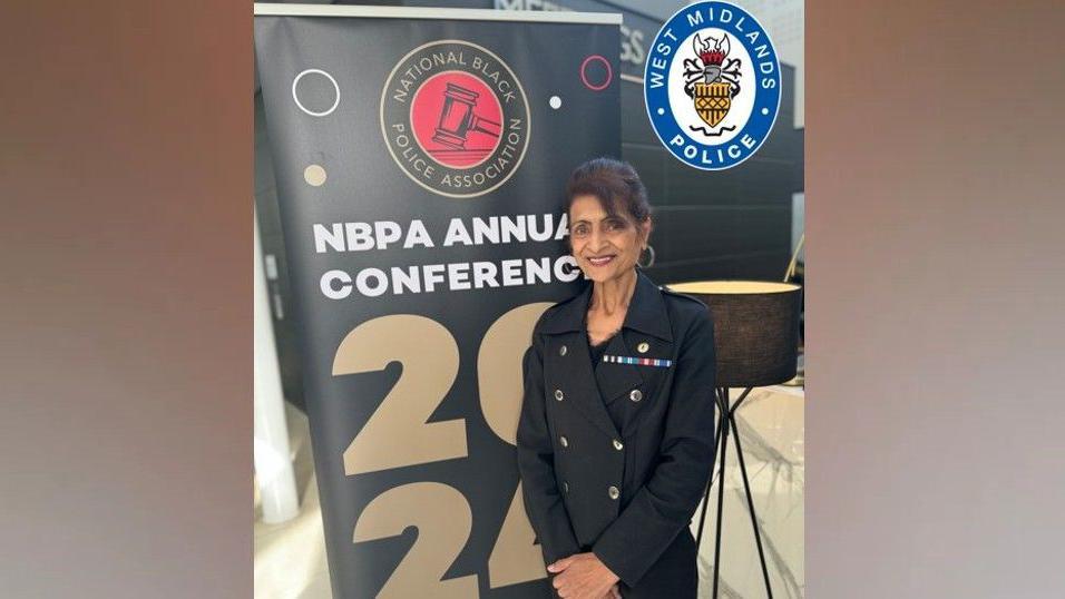 A woman wearing a black blazer jacket and black trousers stands in front of a large black board with text on it which reads "NBPA Annual Conference 2024". She has several embroidered medal ribbons on her jacket.