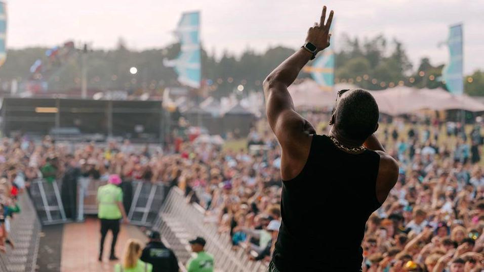 The back view of a performer, with his left arm raised and his head tilted back as he sings. In front of him are crowds held back with a narrow U-shaped barrier. In the background are tents, lights and banners.