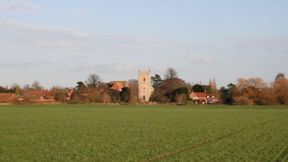 A view of Bassingham village from a ploughed field. There are trees and houses in the distance with a small church in the middle.