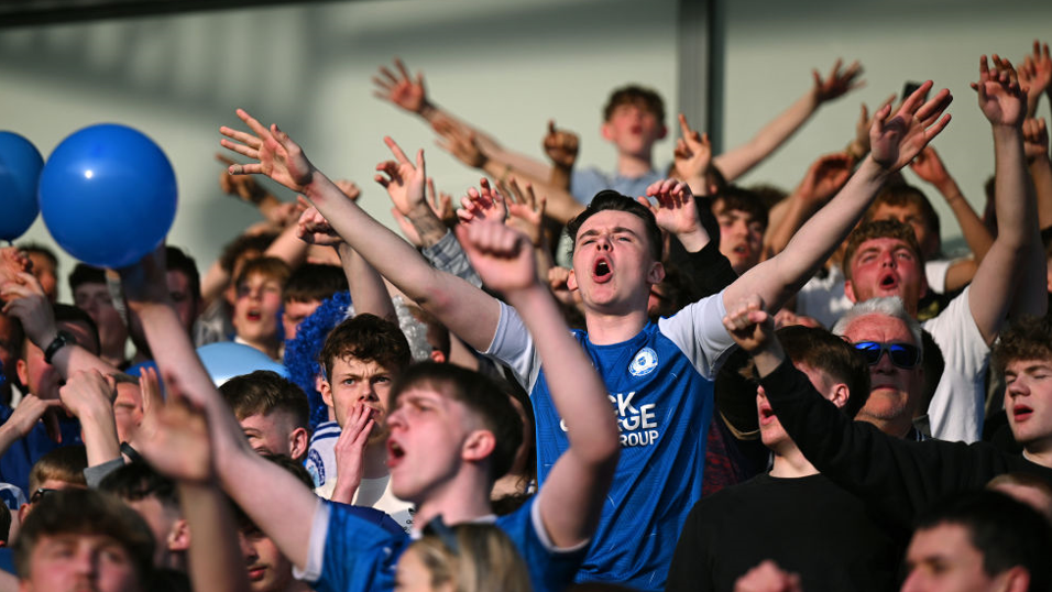 Fans shouting on a football terrace, some with arms aloft.