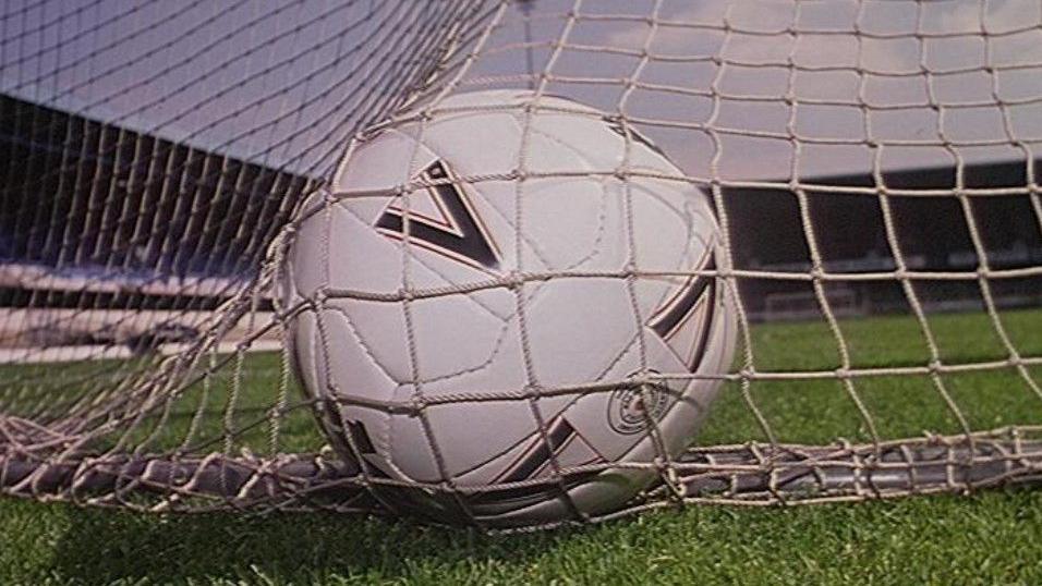 A white football is nestled in a goal net. There are black logos on the ball and green grass all around it. Stadium seating can he seen blurred in the background