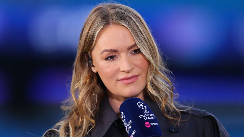 Laura Woods holding a TNT Sports navy microphone while presenting the Champions League. She has wavy blonde hair and is wearing a black coat