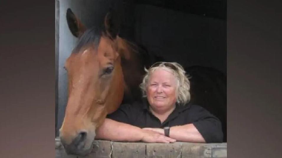 Retired riding instructor Wendy Buckney is stood in a stable next to a horse. She has her arms crossed and has sunglasses atop her blonde hair. She is wearing a black top.