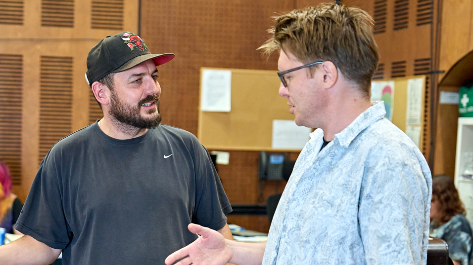 A man in a black t-shirt and a black cap stands talking to a man in glasses and a blue short sleeved shirt. 