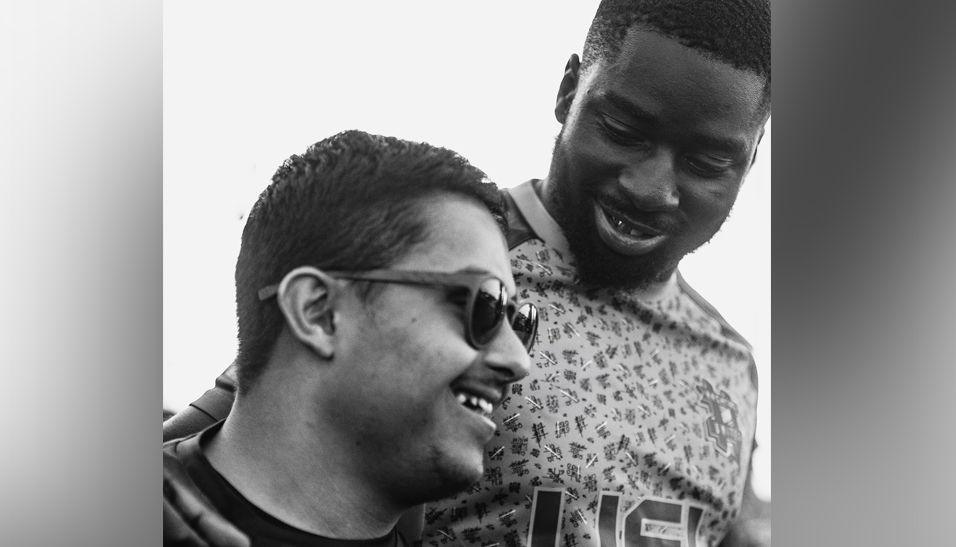 Raman Sidhu is smiling wearing sunglasses side on to the camera. The photo is taken from below. A man in a football shirt has his arm around him, looking down at Raman.