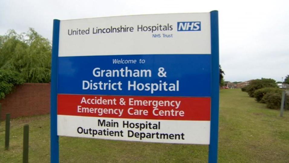 Blue and white Grantham & District Hospital sign on grass. It says United Lincolnshire Hospitals NHS Trust at the top then welcome to Grantham District Hospital with a red A&E sign underneath.