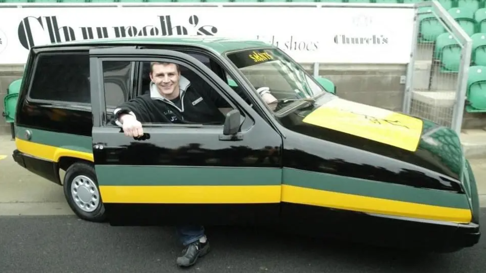 Man sitting in driving seat with door open and leg out - the car is a black Reliant Rialto three-wheeler with green and yellow go stripes in Northampton Saints colours