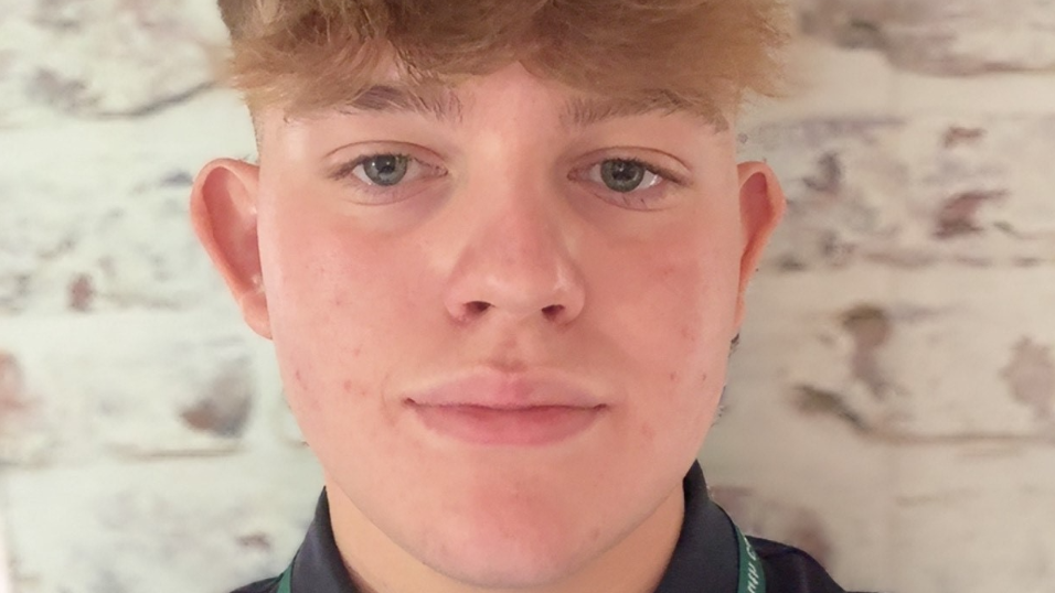 A selfie taken by a young man, smiling at the camera. He has a green lanyard around his neck and a dark polo shirt. He is standing in front of a brick wall, painted white.