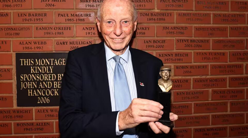 John Rudge is in a suit, standing in front of a brick wall. Each brick has someone's name on it. He is holding a small bronze statue which is a bust of himself