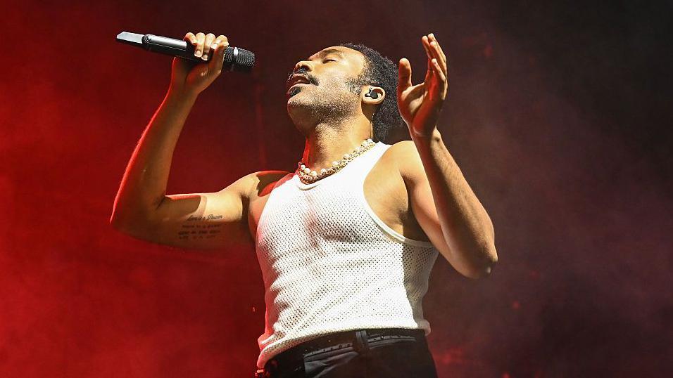 Donald Glover, wearing a white tank top and holding a microphone on stage with smoky red background