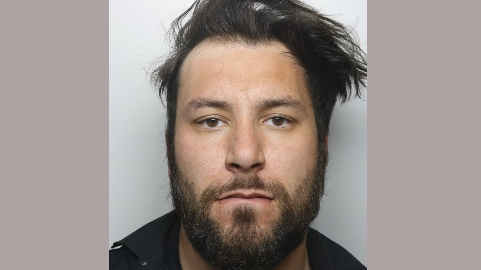 A police custody image of a man with longish black hair, a black beard and brown eyes. He is wearing a dark top. 