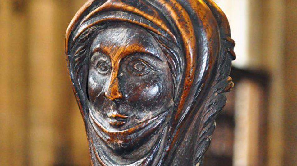 Wood carving of a woman's head. It depicts her wearing a shawl. It is worn and brown in colour.
