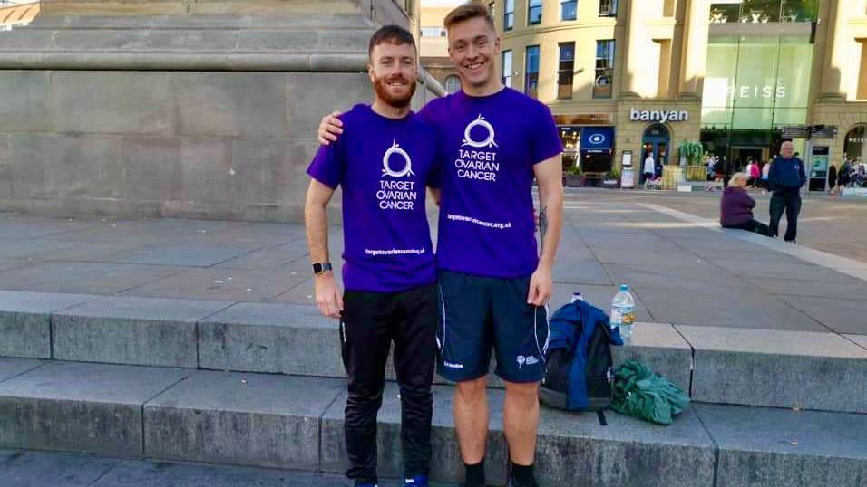 Two men stand in a city centre square with their arms around each other as they pose for a picture. They are both wearing purple t-shirts with the words "Target Ovarian Cancer" in white lettering. A backpack and two bottles of water can be seen on a step behind them.