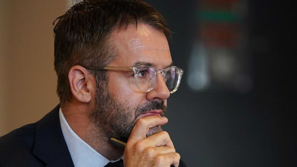 Oliver Coppard, mayor of South Yorkshire, has dark short hair and a trimmed beard and is wearing glasses. He is looking towards the right of the image with his chin resting on his hand