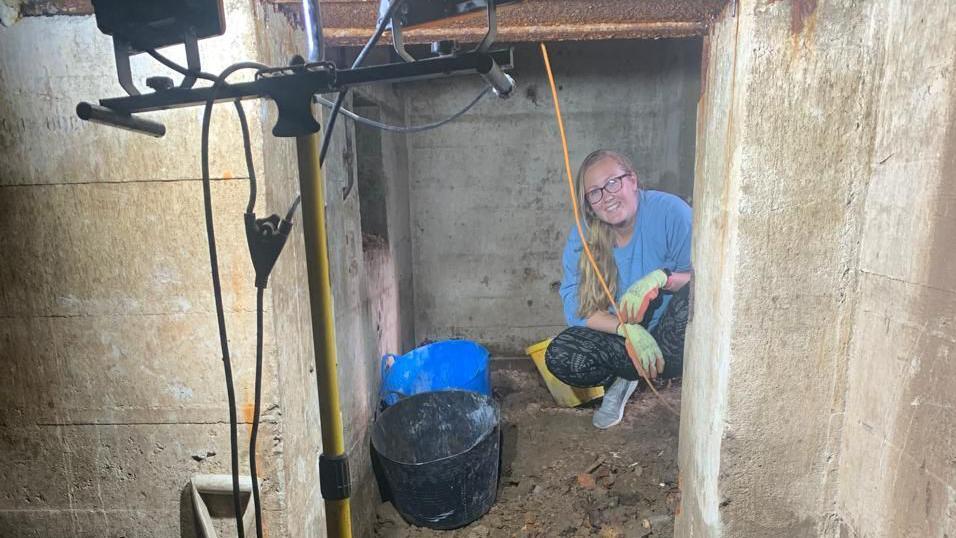 Carrie Tullier squats down inside a German World War Two bunker found underneath her house in Guernsey. She is next to three buckets coloured blue, black and yellow.