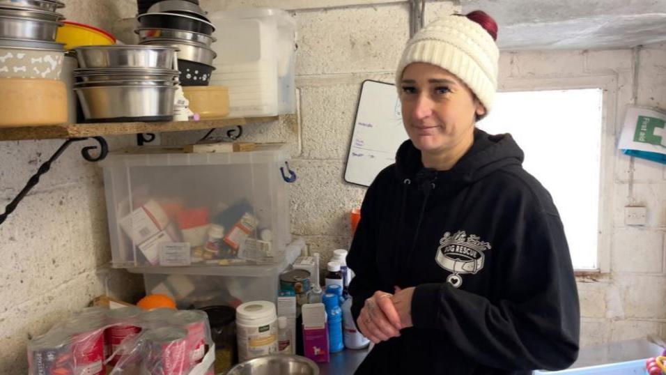 Kymm White, owner of the rescue centre, wearing a dark sweat shirt and white woolly hat looking to camera inside the centre surrounded by medication and dog bowls