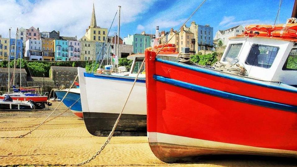 Picture of multicoloured houses in Tenby, overlooking the harbour at low tide. Several boats can be seen moored on the sand.