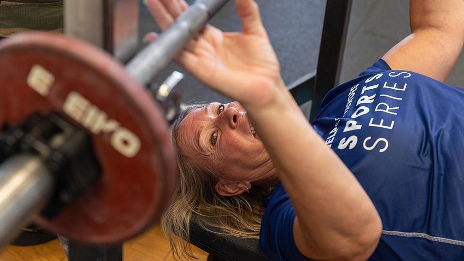 Shirley Ghobrial with blond hair wearing a blue t-shirt with the words Help the Heroes Sports Series in white printed on it attempts to a power lift.