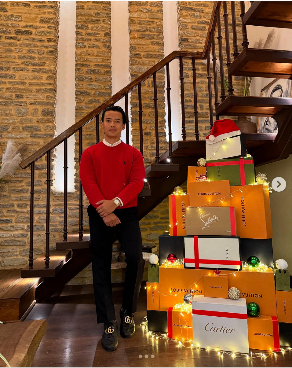 Saugat Thapa, in a red sweater and black trousers, stands next to a Christmas tree made of boxes of luxury brands 