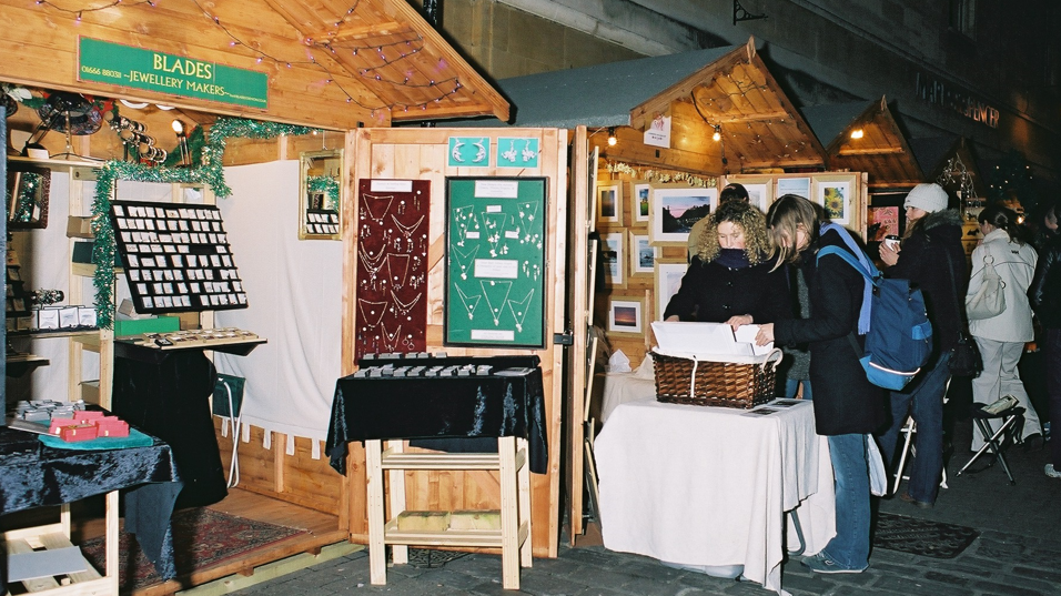 A line of chalets with the nearest one selling jewellery. Two women are looking at a basket of prints at another stall selling paintings.