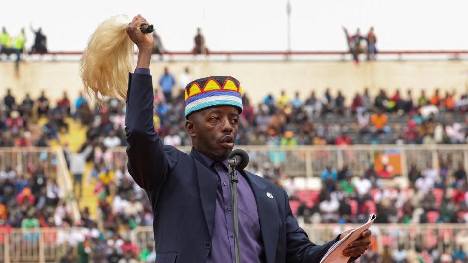 Raila Odinga Junior waiving a fly whisk while wearing atraditional cap