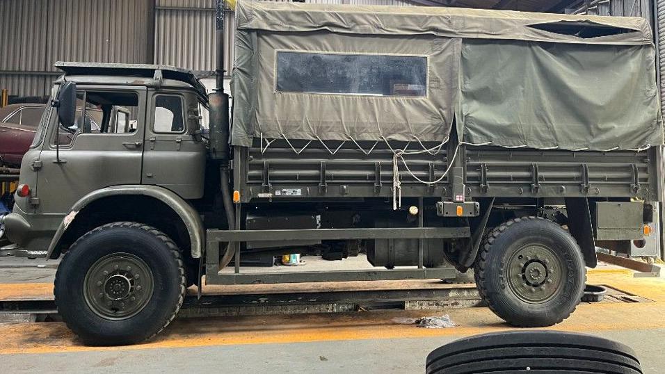 The side-profile of the green military vehicle, which has green tarpaulin covering the area for storage