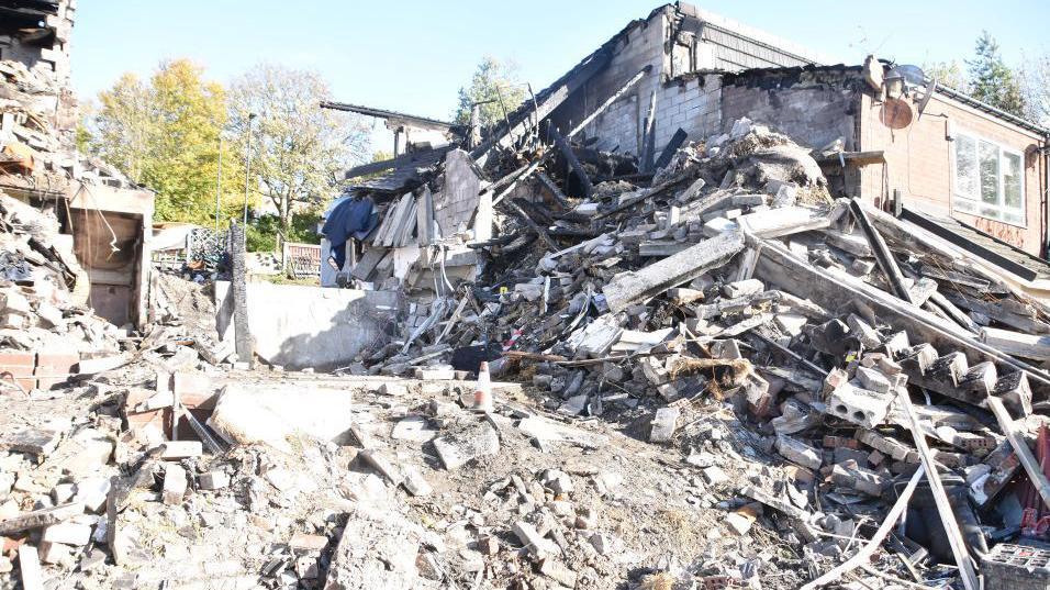 A pile of masonry rubble, bricks and large pieces of concrete, where a house used to be. Another house is just beyond, attached to the rubble.