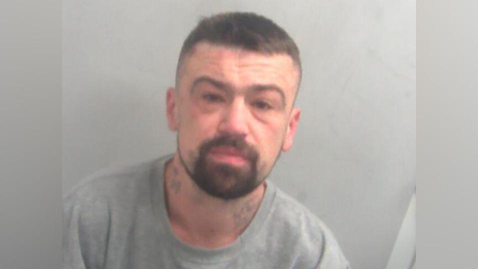 A custody image of a man. He has a brown beard and he is wearing a grey shirt.