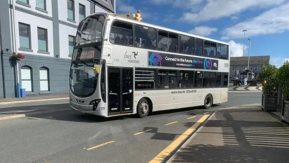 A silver double-decker bus drives on a road next to a roundabout and large building.