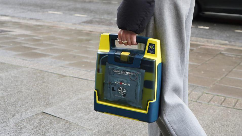 A close up of a defibrillator being carried down a London street. The device is in a yellow and dark blue plastic case with a see through panel at the front. 