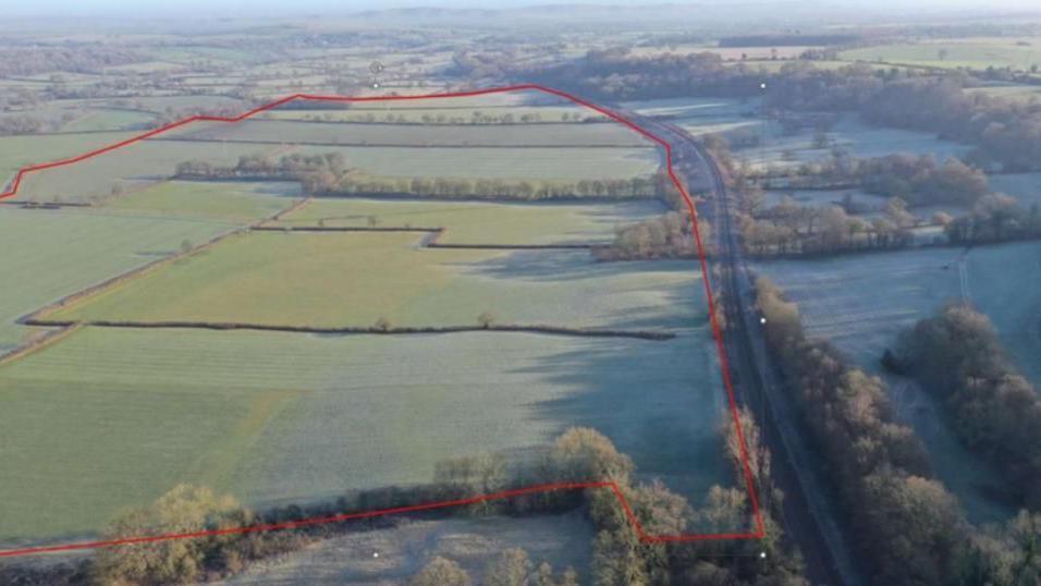 A drone shot of fields and countryside, a large red line drawn round the fields where solar panels would be