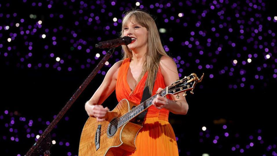 Taylor Swift in an orange dress, holding a guitar, sings on stage.