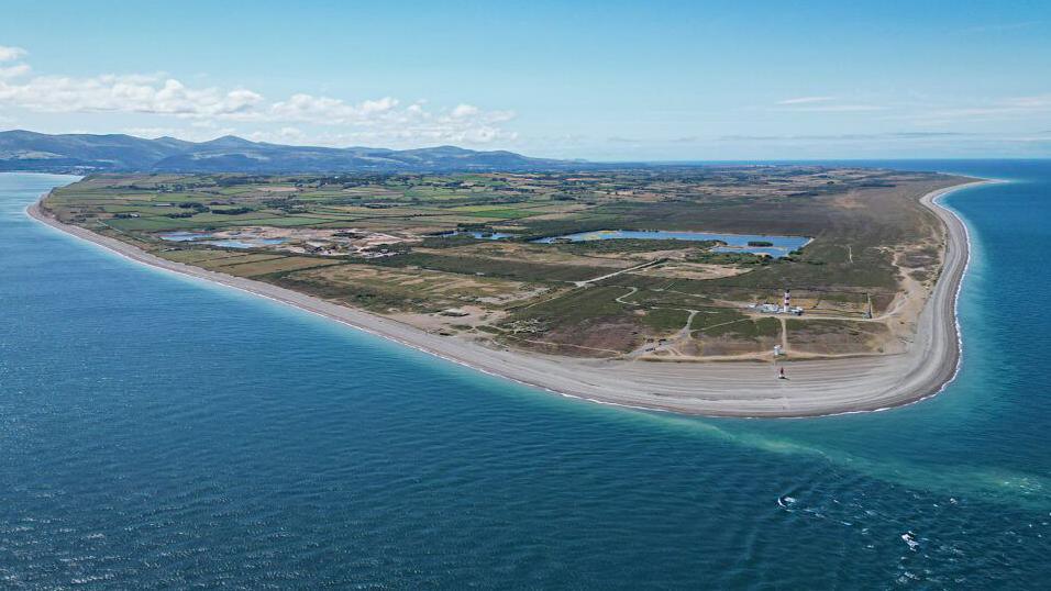 An aerial view of the northern tip of the Isle of Man. There green fields edged by a stretch of sandy beach beach that meets the sea, which is a vibrant turquoise colour.