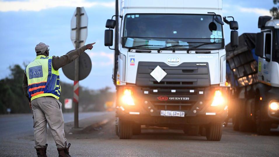 A traffic officer, with his back to the camera, directs a truck with its lights on waiting to cross South Africa's border into Mozambique - Komatipoort, South Africa, July 2023