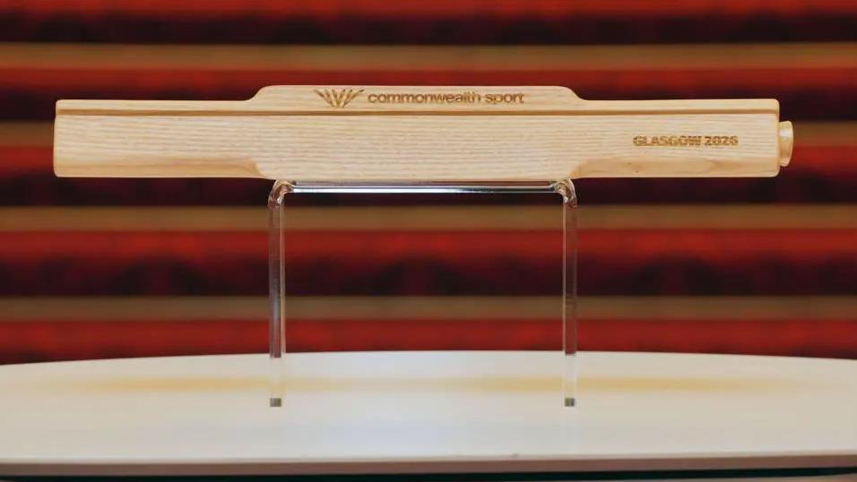 A brown wooden baton is placed on a clear stand in front of a red background. The words commonwealth sport and glasgow 2026 have been etched into it.