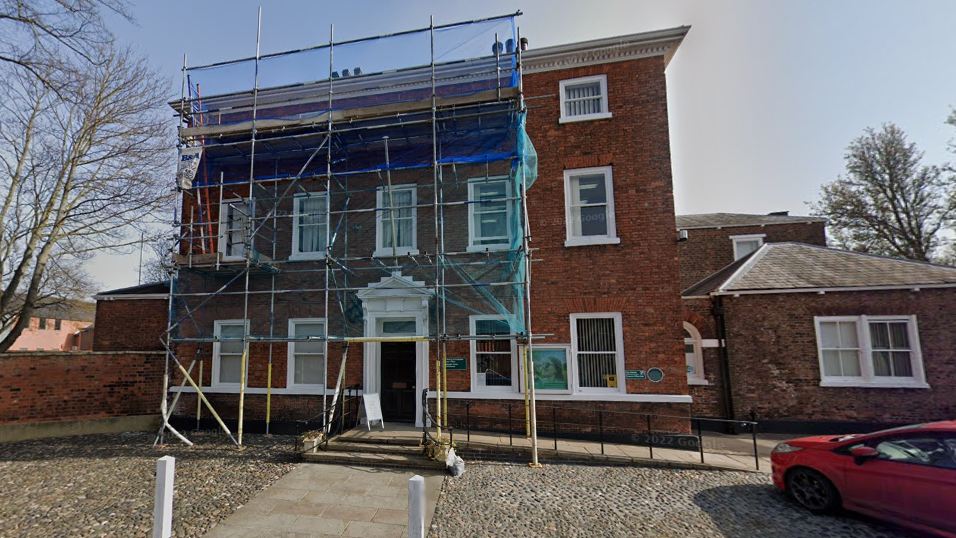 A large red-brick building with a black front door and cobbles to the front, it has scaffolding up on the exterior. 