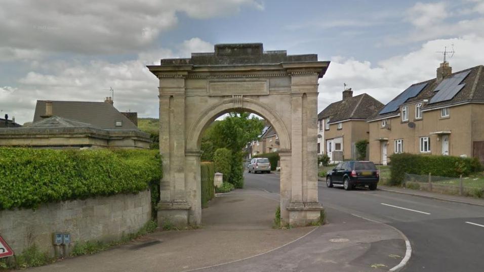 The Anti-Slavery Arch in Paganhill. It is a large rectangular stone archway on the site of a curved pavement, next to the road. There is a row of semi-detached houses on the opposite side of the road. Beside the archway is a low wall and hedge. 