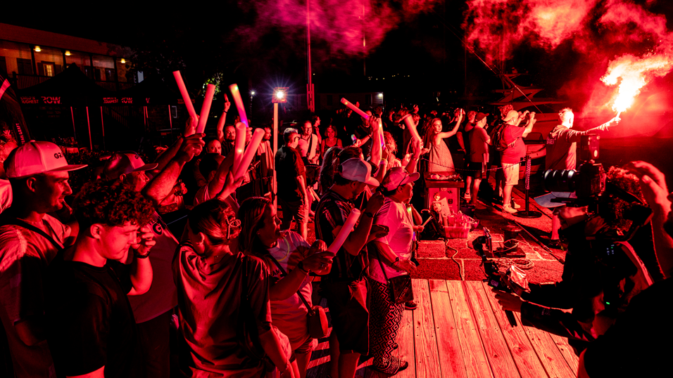 A crowd of people cheer and wave as the boat with the four men who made up Team Graft pull into the pontoon. The scene is lit by red flares.