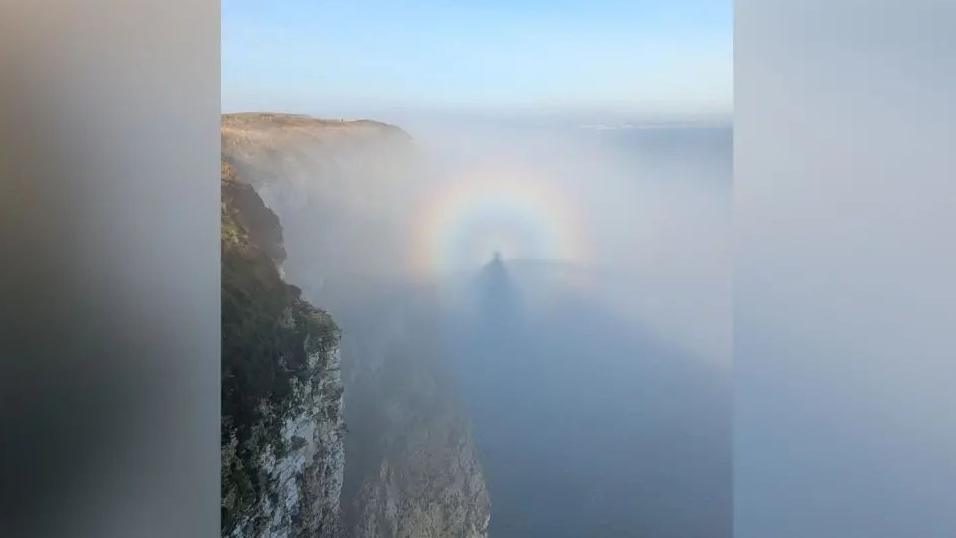 A Brocken Spectre - an image of a person's shadow captured in the mist. It has a circular rainbow surrounding it and the cliffs and sea can be seen in the image.