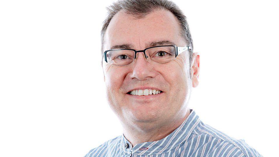 A headshot of Stephen Foster. He is wearing glasses and a striped shirt. He is smiling into the camera while standing in front of a white backdrop.
