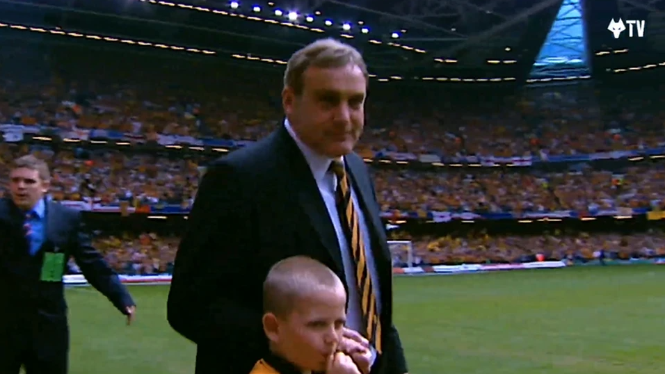 Mitch as a boy is sucking his thumb and holding the hand of a man who has dark hair and is wearing a dark suit, blue shirt and gold and black striped tie, as they walk out at the start of a football match. There are crowds in the stands surrounding the green pitch. 