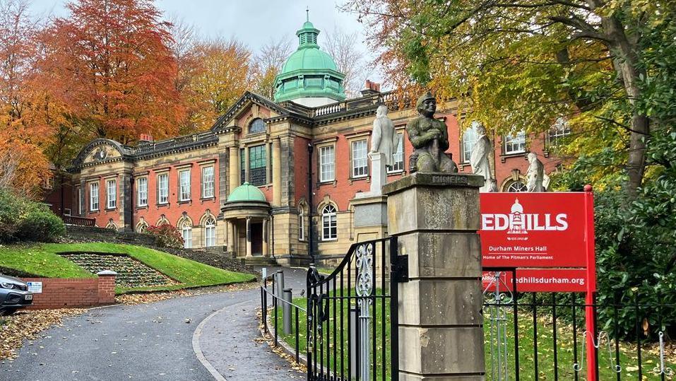 The outside of Redhills which is a large beige and orange stone building with a bright green dome. Outside are a few white statues facing the driveway in front. There is a red sign at the front which reads 'Redhills' next to a black metal gate.
There are trees surrounding the building which have orange, green and red leaves.