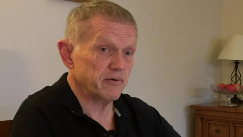 Richard Barker, who has short fair hair and is wearing a black polo shirt, sits in his living room, speaking to a BBC reporter who is out of shot